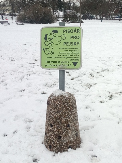 Un cartello verde nel parco, con la scritta «Pisoár pro pejsky» e il disegno di un cane che tiene in bocca un osso gigante.