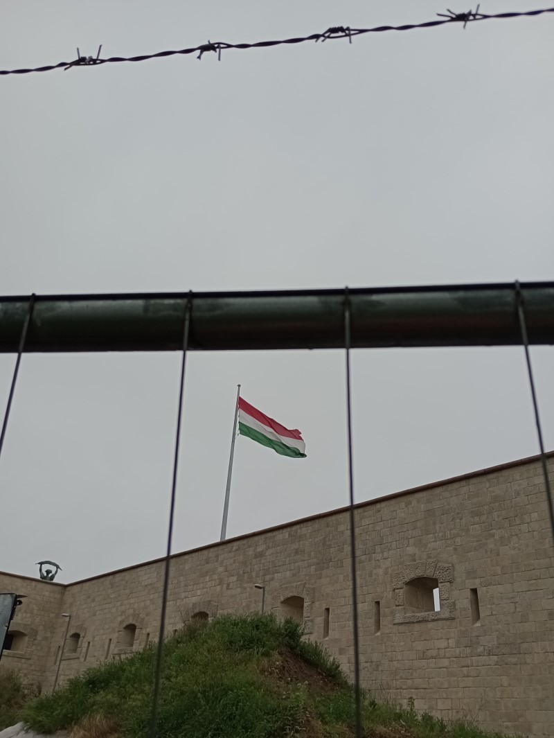 Bandiera ungherese sventolante su un pennone vista da dietro le transenne e il filo spinato.