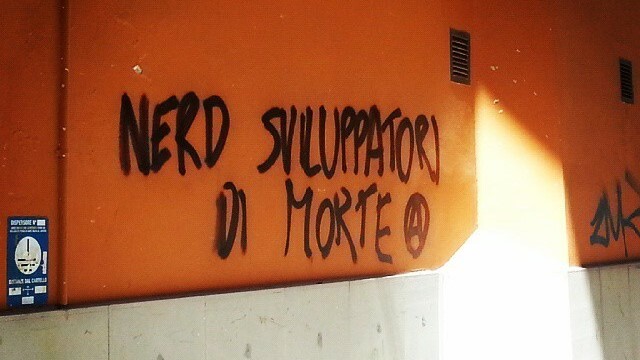 Una scritta nera su un muro arancione: “NERD SVILUPPATORI DI MORTE”, seguita da una “A” cerchiata.