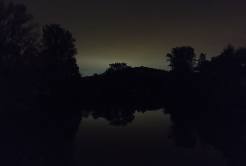 A hill in almost-complete darkness, reflected in the still waters of a river. Behind the hill, an unnatural light glows in the sky.