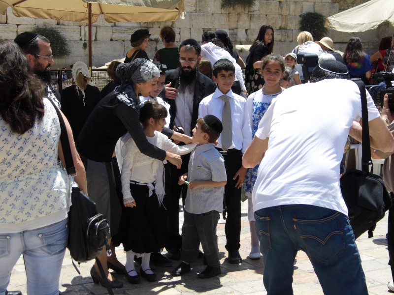 On the foreground of the Western Wall, a family of neatly-dressed Orthodox Jews: father, mother, five children aged about six to thirteen; an uncle overlooks, a man takes their picture.