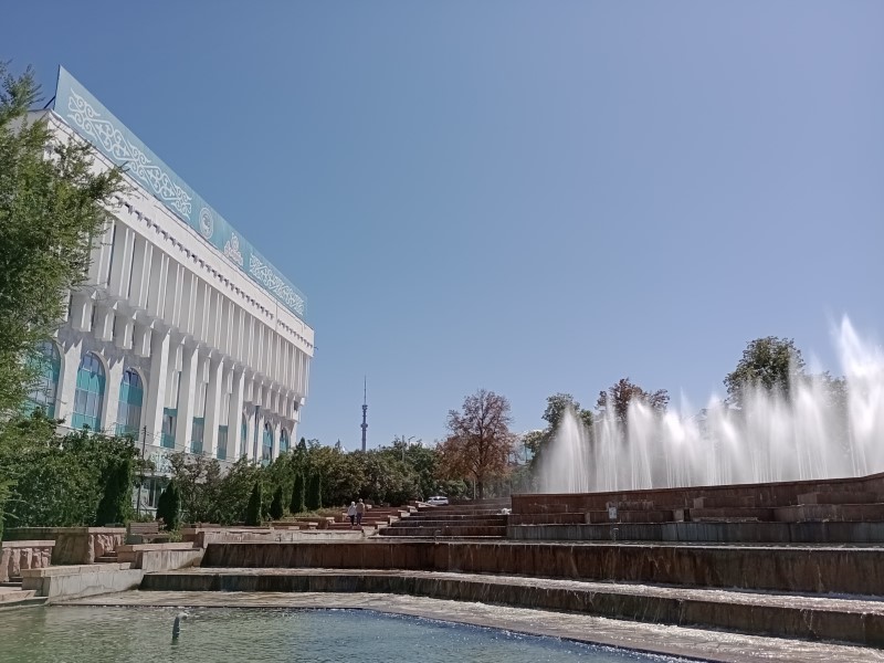 Sulla sinistra, un edificio con facciata in cemento e vetro, sormontato da un’insegna azzurra con il medesimo motivo grafico della bandiera nazionale. Sulla destra, una fontana a scalinata. Lontane sullo sfondo, una torre e delle montagne innevate.